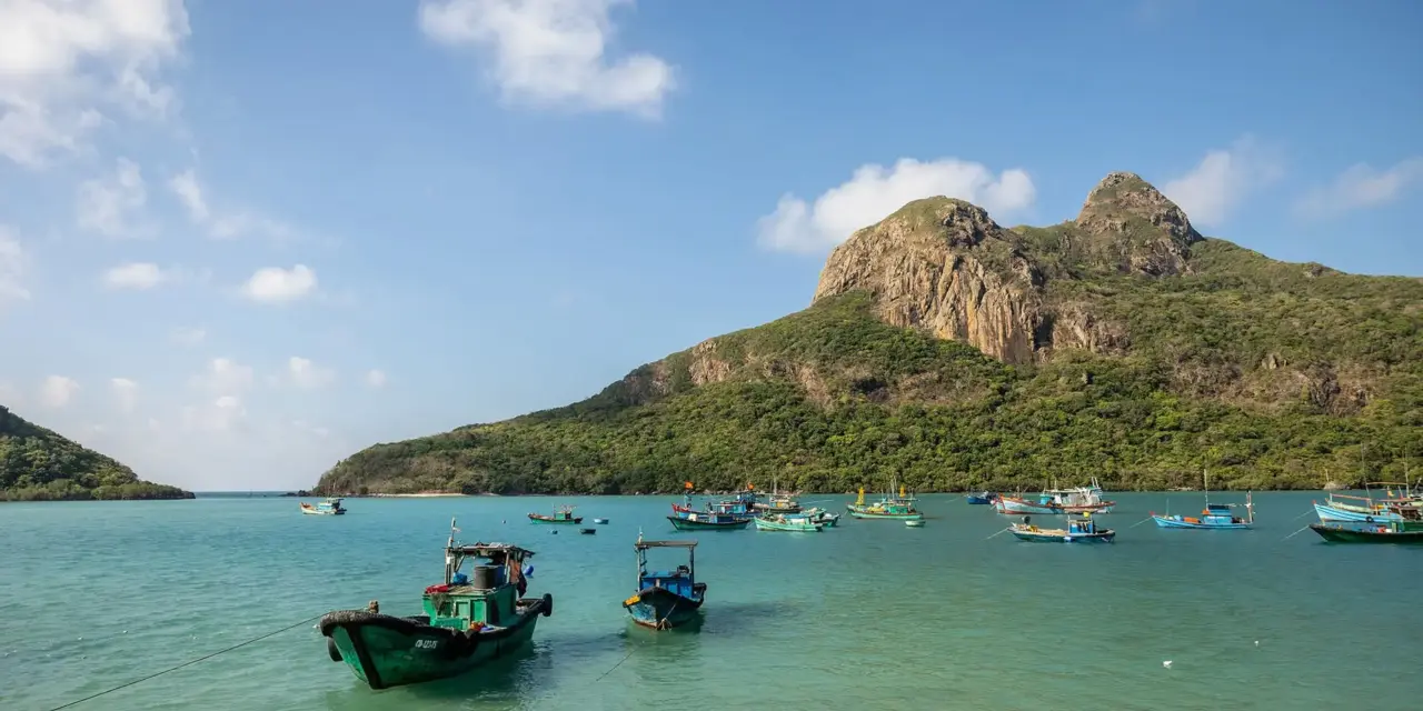 Thuyền đánh cá neo trên vịnh nước xanh, phía sau là ngọn núi đá phủ rừng