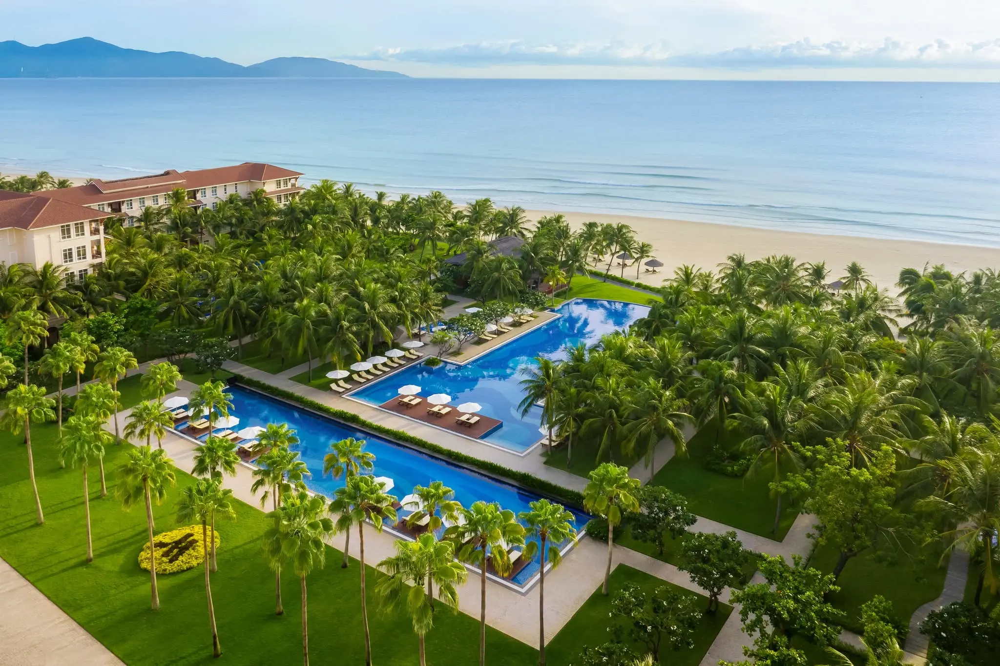 Hồ bơi dài giữa vườn cọ của một resort sát bãi biển, phía xa là biển và dãy núi