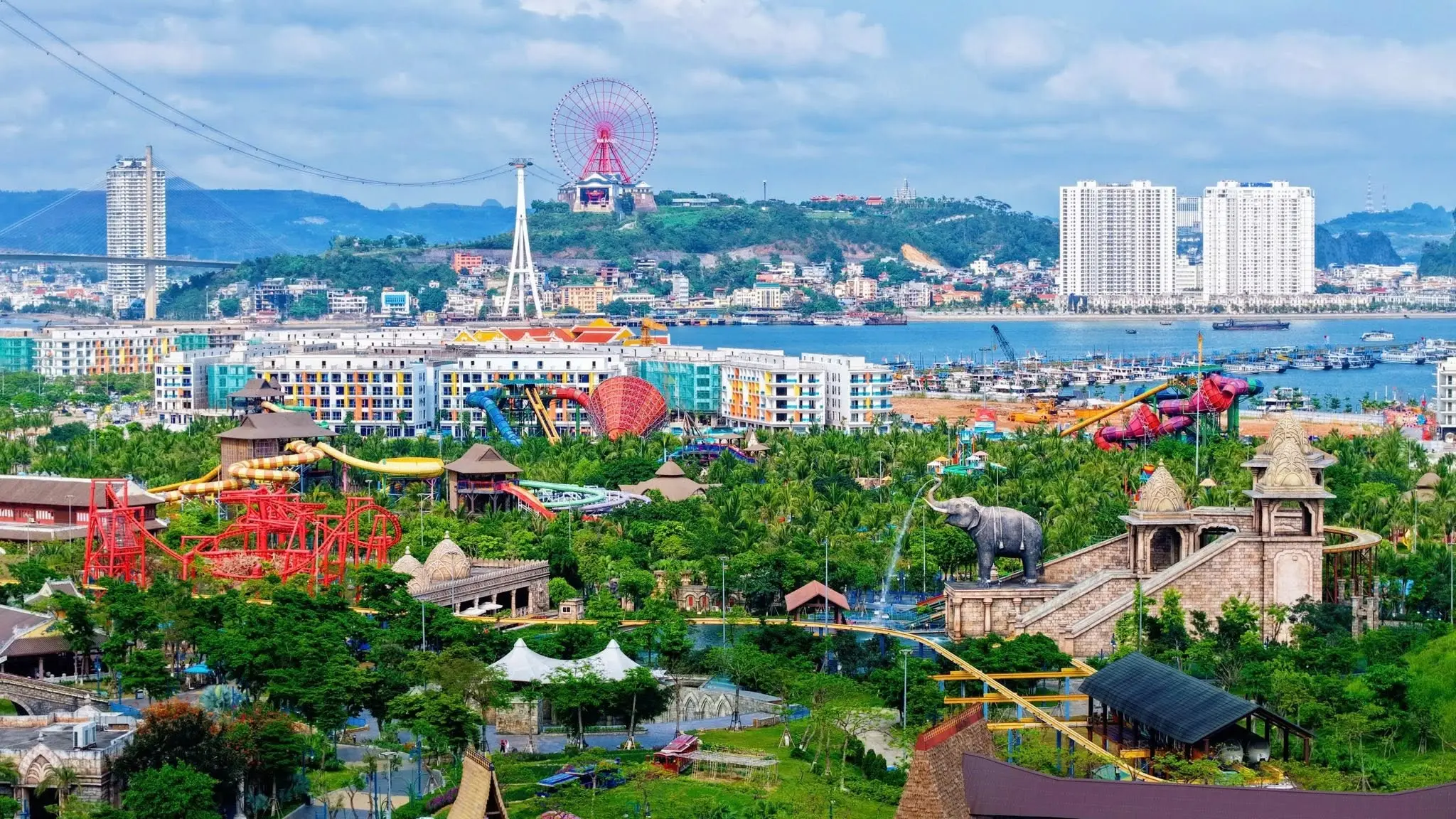Khu vui chơi nhiều màu sắc với máng trượt nước và tượng voi, phía xa là vòng quay và nhà cao tầng ven vịnh
