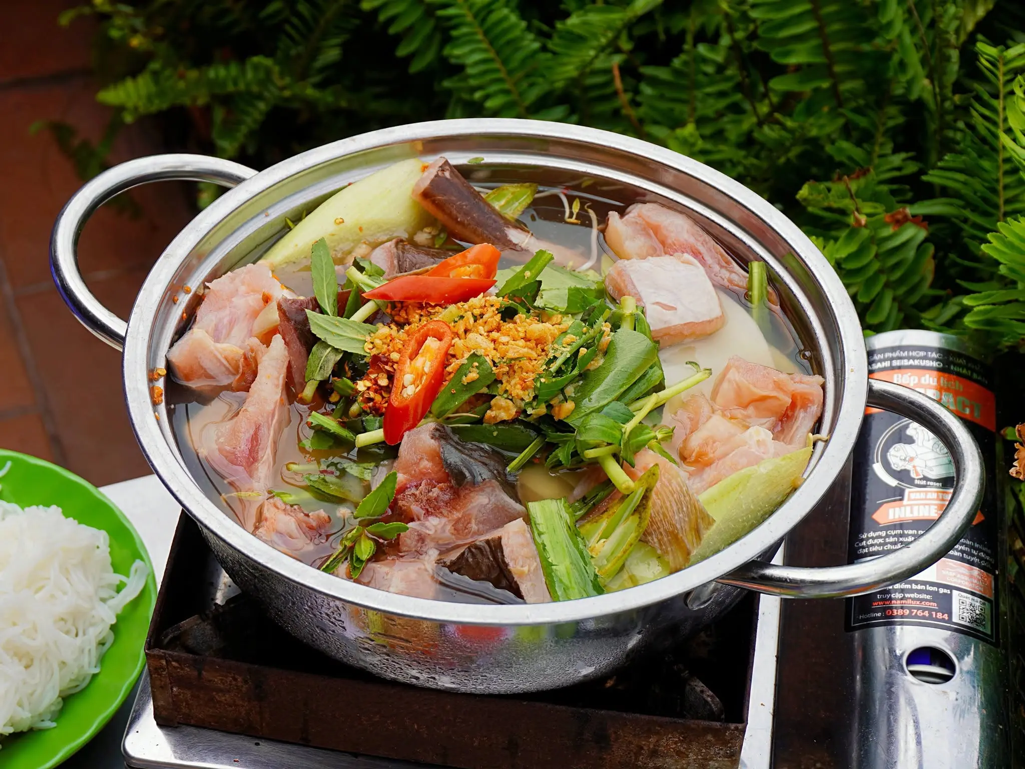 Nồi lẩu inox với cá, rau thơm và ớt đặt trên bếp ga mini, bên cạnh là đĩa bún