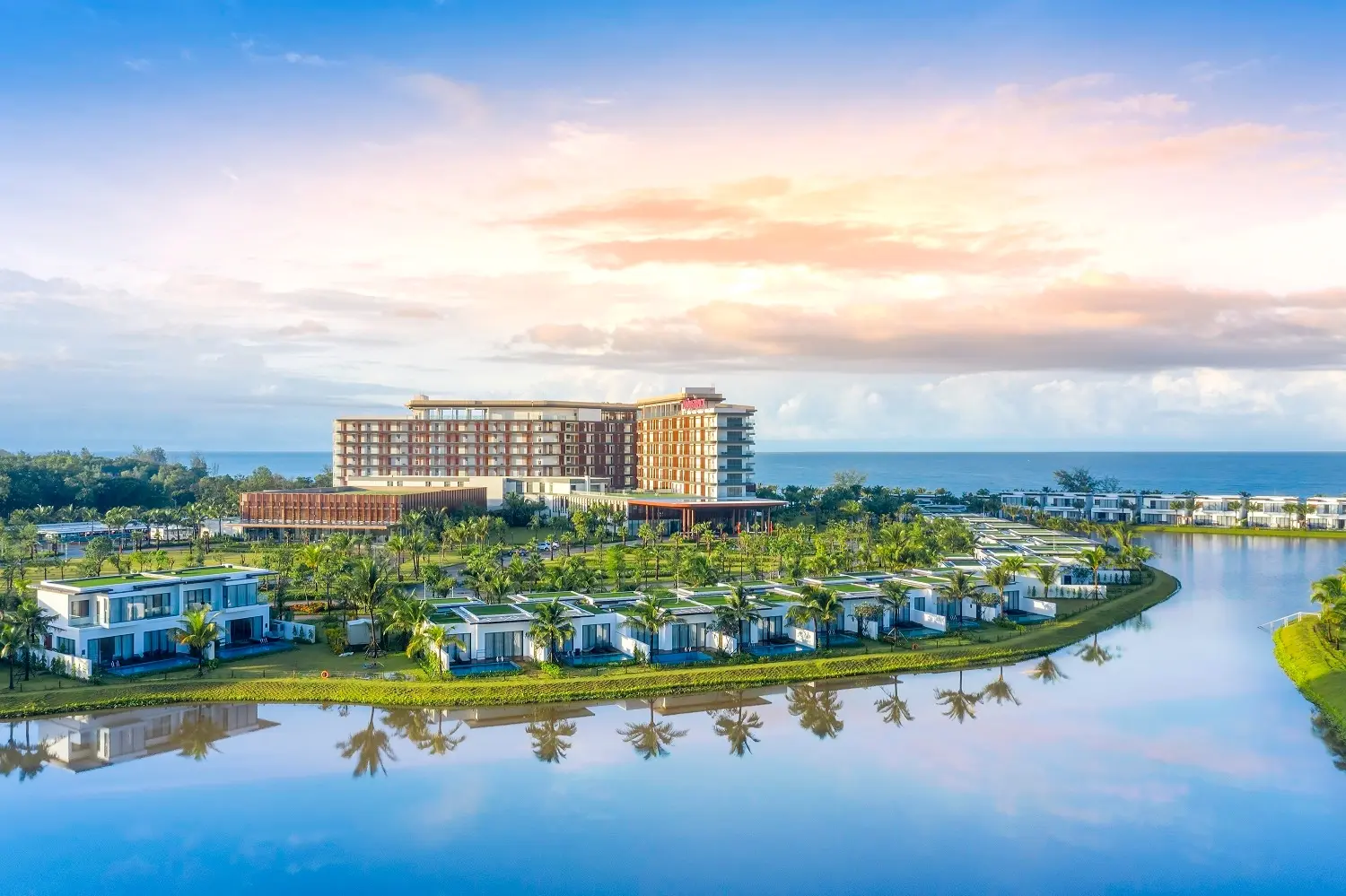 Khu biệt thự resort bên mặt hồ phẳng lặng, phía sau là khách sạn cao tầng sát biển lúc bình minh