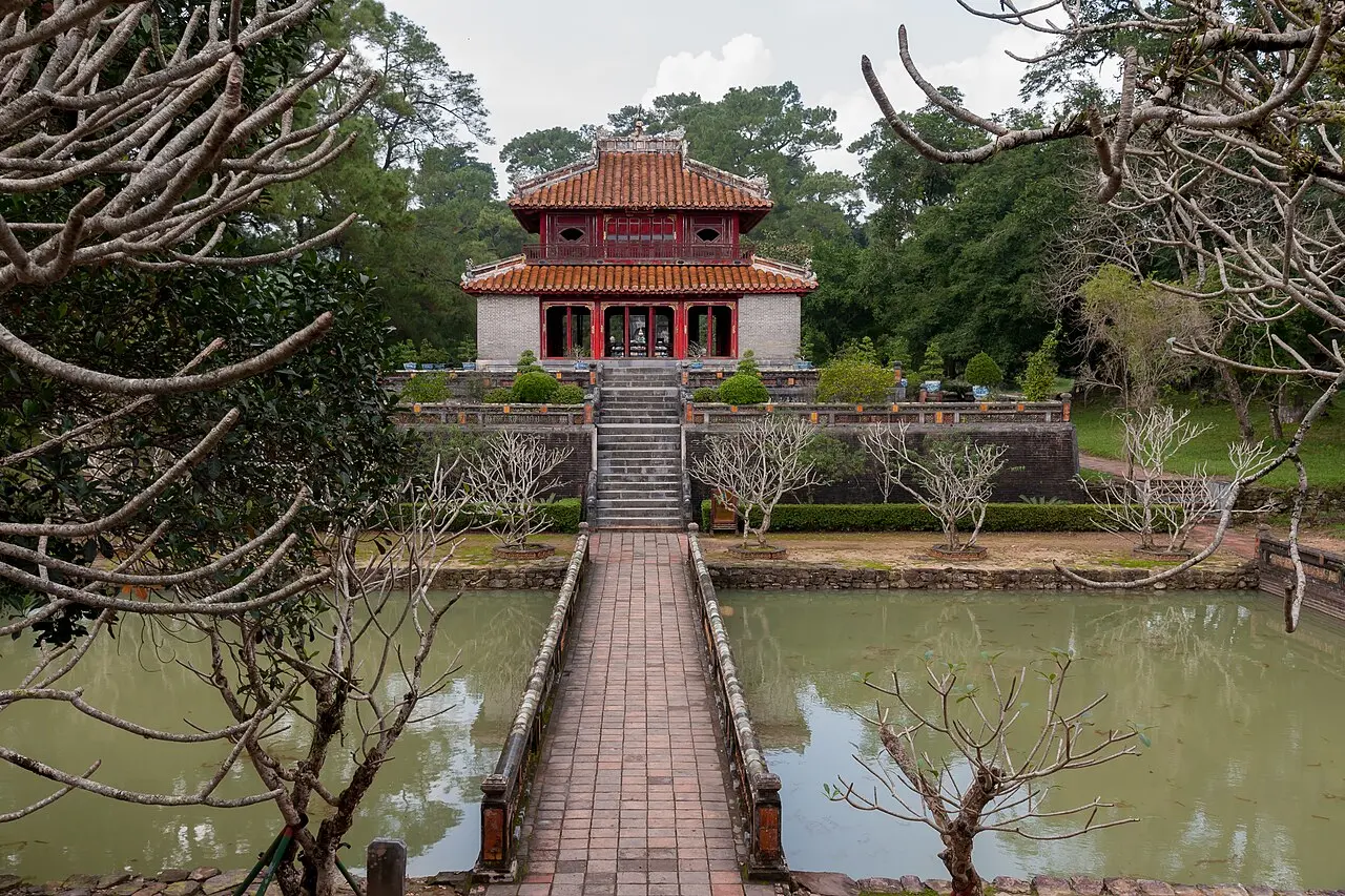 Cây cầu lát gạch bắc qua hồ dẫn tới lầu hai tầng mái ngói đỏ giữa vườn cây